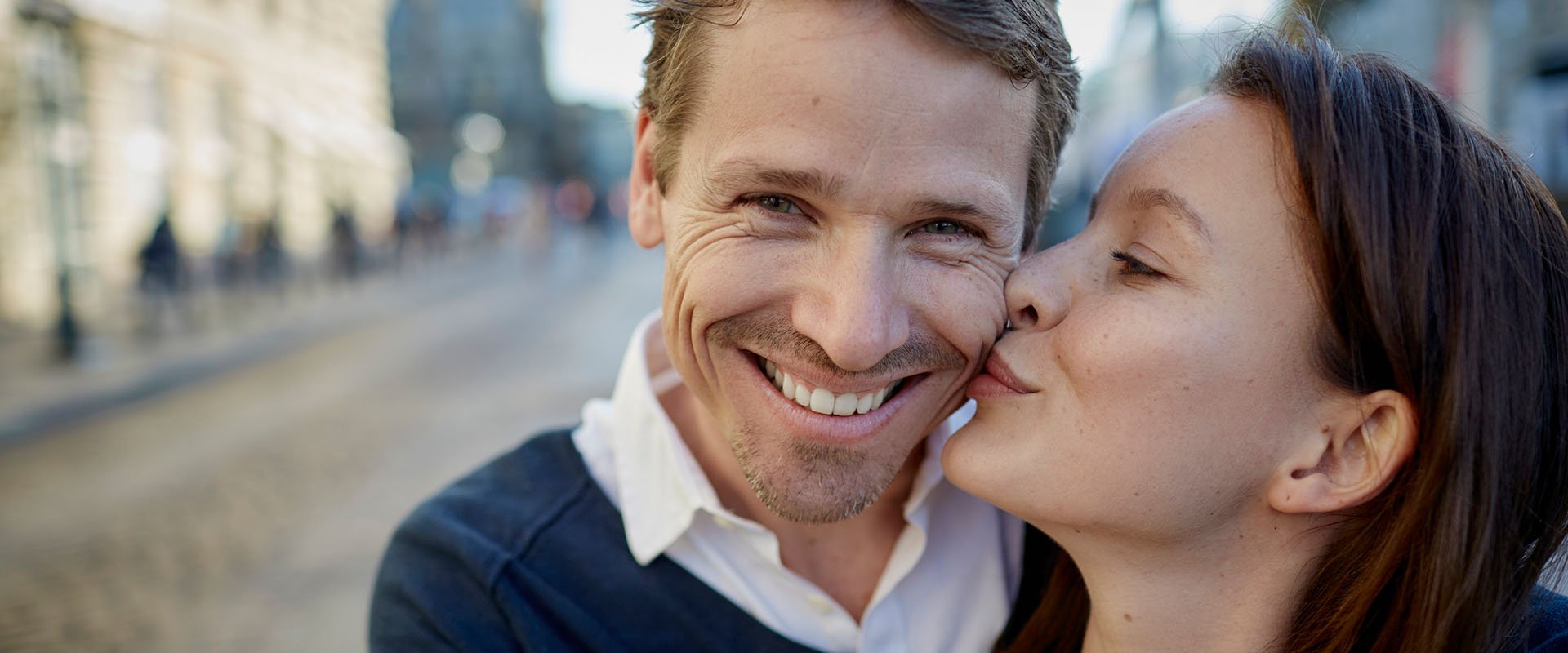 Eine Frau küsst ihren zufrieden lächelnden Mann auf der Straße