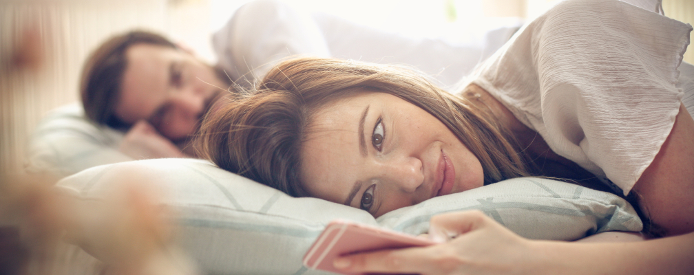 Ein Pärchen liegt im Bett. Die Frau dreht sich vom Mann weg und schaut lächelnd auf ihr Smartphone.